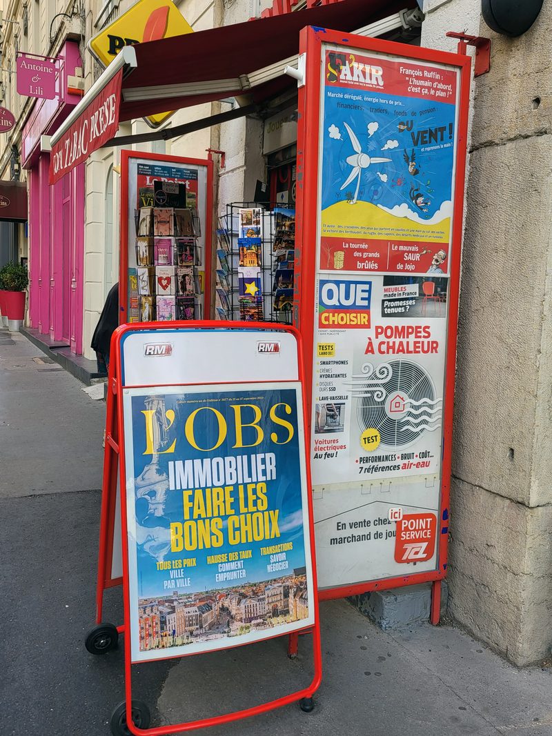 Chevalet sur trottoir et panneau vitré devant un tabac-presse