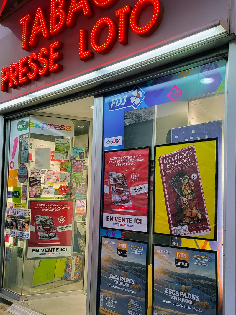 Devanture de tabac-presse éclairée en soirée, affiches visibles depuis la rue
