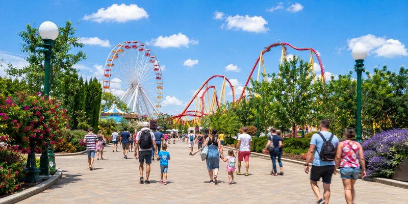 Allée d'un parc d'attractions en plein jour, grande roue et montagnes russes en arrière-plan